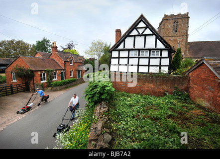 Gärtner schieben Rasenmäher durch das Dorf Redmarley D'Abitot, Gloucestershire mit der Kirche von St. Bartholomäus Stockfoto