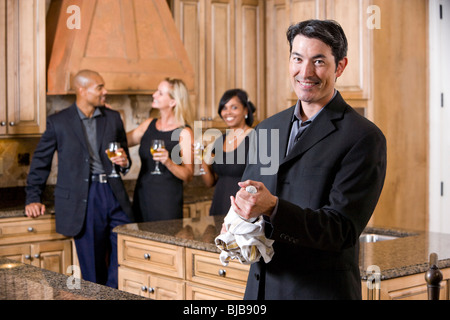 Mann mit Champagner-Flasche in Küche, Gruppe von Freunden im Chat im Hintergrund Stockfoto