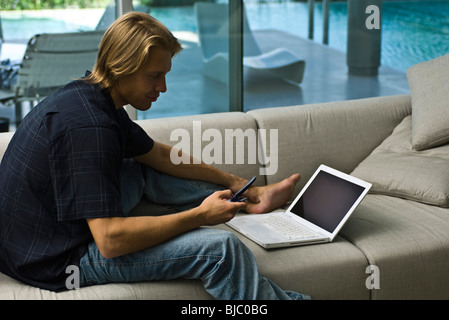 Mann, sitzend auf Sofa mit Laptop in der Nähe, mit Handy Stockfoto