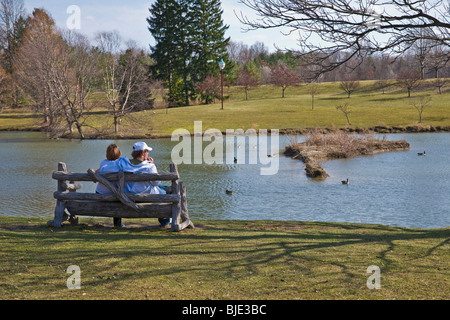 Ein junges Paar sitzt auf einer Holzbank im Stadtpark und beobachtet Gänse, die in einem Teich in Ohio, USA, schwimmen Stockfoto