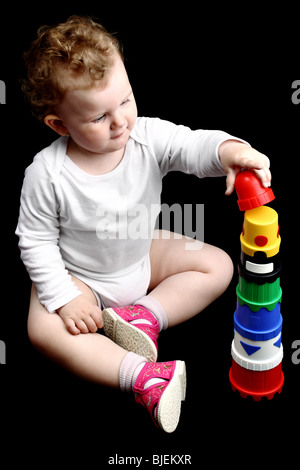 Lockige Baby sitzen auf dem Boden einen Turm zu bauen, mit Stapeln Verschachtelung Tassen isoliert auf schwarz Stockfoto