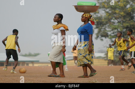 Frauen fahren Sie mit ihrer täglichen Arbeit die Güter transportieren, wie sie Kinder Fußball spielen passieren. Kumasi, Ghana, Westafrika, Afrika Stockfoto