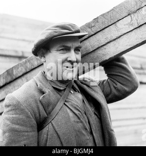 England, 1950er Jahre. Nahaufnahme eines Arbeitnehmers für London Brick Company, tragen eine Jacke und Mütze, tragen drei Holzplatten. Stockfoto