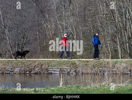 Senior paar einen Mann eine Frau ihren Hund entlang des Kanals in einem Park spazieren gehen. Zeitigen Frühjahr Tag tragen sie bunte Jacken. Stockfoto