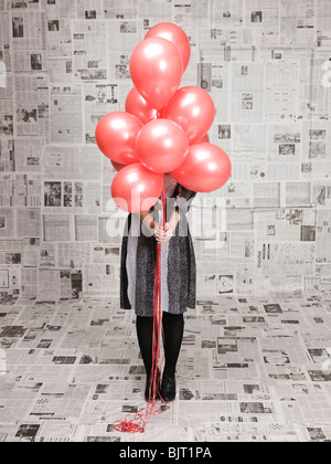 Junge Frau Gesicht hinter Haufen Luftballons in Zeitungen versteckt abgedeckt Zimmer, Studio gedreht Stockfoto