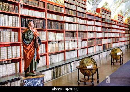 PRAG, Tschechische Republik — das kunstvolle Innere der Strahov-Bibliothek, Teil des Strahov-Klosters in Prag. Auf der linken Seite steht eine Statue des heiligen Johannes des Evangelisten, auf der rechten Seite ein speziell entworfenes Buchrad, eine innovative Erfindung aus der Renaissance, die es den Lesern ermöglicht, mehrere Bücher gleichzeitig durch Drehen des Rades zu referenzieren. Die aufwendige barocke Einrichtung der Bibliothek, einschließlich Freskendecken und vergoldeten Bücherregalen, zeigt ihre Bedeutung als Wissensspeicher und kulturelles Erbe. Stockfoto