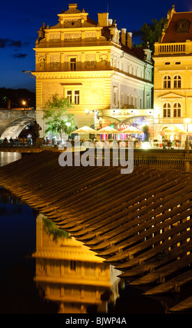 Bedřich Smetana Museum und Klub Lavka an der Moldau Prag Tschechische Republik // PRAG, Tschechische Republik — das beleuchtete Bedřich Smetana Museum und der benachbarte Klub Lávka spiegeln sich in der Moldau neben der Karlsbrücke in der Altstadt. Das Museum ist dem berühmten tschechischen Komponisten gewidmet und befindet sich im ehemaligen Gebäude der Altstadt, einem Gebäude der Neorenaissance, das 1884 fertiggestellt wurde. Im Vordergrund sind historische Holzeisbrecher (ledolamy) entworfen, um die Säulen der nahegelegenen Karlsbrücke zu schützen, die teilweise im Hintergrund sichtbar ist. Das Museum ist ein B Stockfoto