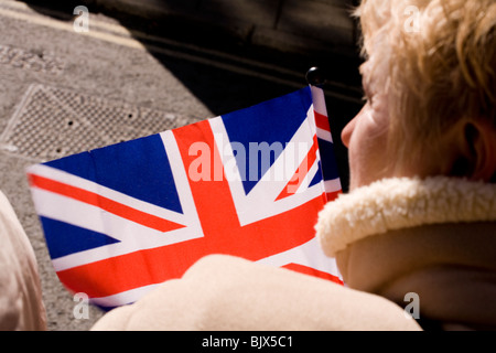 Eine Frau Union Jack Flagge weht im Wind wie sie mit Spannung das Aussehen der Queen erwartet. Stockfoto