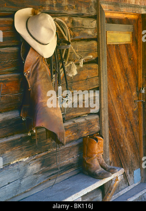 Cowboy-Hut, Chaps, Tack und Seil hängen auf einer Blockwand mit Cowboy-Stiefel auf Holzbank, Montana, USA Stockfoto