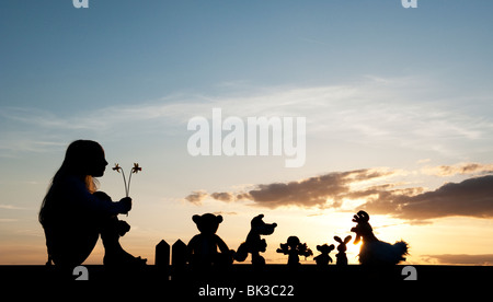 Junges Mädchen mit Narzissen und eine Stoffpuppe, Huhn, Fuchs, Hase und Bär Stofftiere sitzen auf ein Tor bei Sonnenuntergang. Silhouette Stockfoto