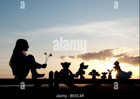 Junge Mädchen mit Narzissen und eine Stoffpuppe, Huhn, Fuchs, Hase und Bär Stofftiere sitzen auf ein Tor bei Sonnenuntergang. Silhouette Stockfoto