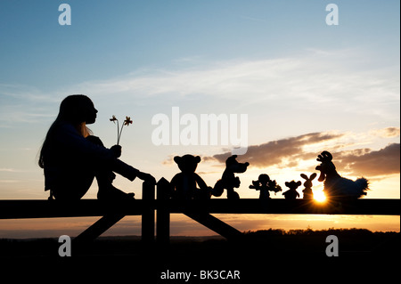 Junge Mädchen mit Narzissen und eine Stoffpuppe, Huhn, Fuchs, Hase und Bär Stofftiere sitzen auf ein Tor bei Sonnenuntergang. Silhouette Stockfoto