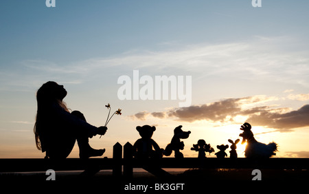 Junge Mädchen mit Narzissen und eine Stoffpuppe, Huhn, Fuchs, Hase und Bär Stofftiere sitzen auf ein Tor bei Sonnenuntergang. Silhouette Stockfoto