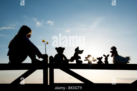 Junge Mädchen mit Narzissen und eine Stoffpuppe, Huhn, Fuchs, Hase und Bär Stofftiere sitzen auf ein Tor bei Sonnenuntergang. Silhouette Stockfoto