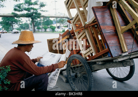 Mann liest eine Zeitung und trägt einen Strohhut neben seinem Wagen mit Holzmöbeln in Bangkok, Thailand Stockfoto