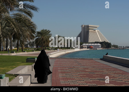 Arabische Frau zu Fuß entlang der Corniche mit dem Sheraton Hotel im Hintergrund, Doha, Katar Stockfoto