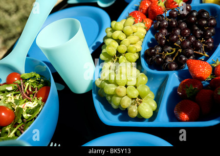 Im Freien essen oder ein Picknick auf einem schwarzen Tisch mit blauen Kunststoff Schüsseln, Tassen und Krüge mit Obst und Salat Stockfoto