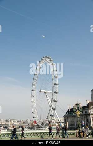 Touristen überqueren Westminster Brücke, vorbei am London Eye und altes Rathaus. Ein Jet geht über dem Kopf. Stockfoto