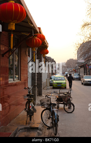 Sonnenuntergang in Peking Hutong mit Fahrrädern und Autos parkten auf einer engen Gasse Straße vor einem Restaurant mit roten Laternen. Stockfoto