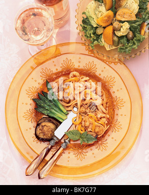 Fettucine mit Garnelen, Spargel und Auberginen auf gold Relief Glasplatten, hell rosa Stoff, Salat mit gelben Tomaten, Oliven Stockfoto