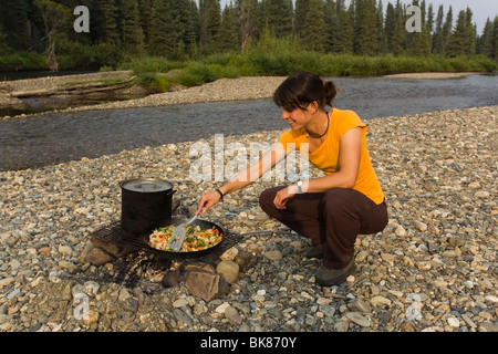 Junge Frau, Kochen, Braten Huhn rühren braten am Lagerfeuer, Kiesbank, oben Liard River, Yukon Territorium, Kanada Stockfoto