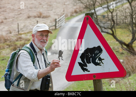 Ein Mann hält eine Kröte, die von einem Auto neben einer Kröte überquert Warnschild auf einer Straße in der Nähe von Aktien Reservoir in Lancashire, UK abgeflacht. Stockfoto