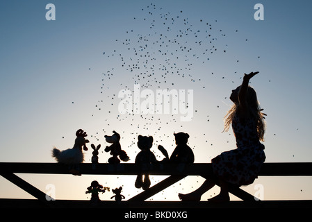 Mädchen Wurfsterne über eine Stoffpuppe, Huhn, Fuchs, Hase und Teddy Bear Stofftiere sitzen auf ein Tor bei Sonnenuntergang. Silhouette Stockfoto