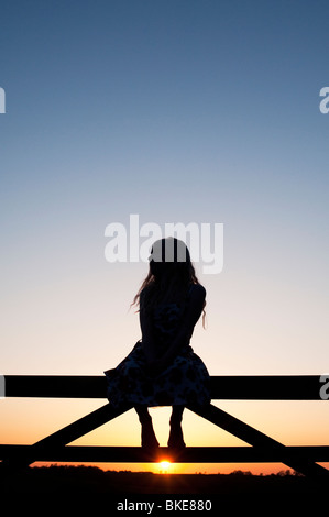 Junges Mädchen sitzt auf einem Tor bei Sonnenuntergang. Silhouette Stockfoto