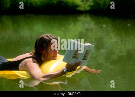 Junge Frau mit Buch beim Schwimmen in ländlichen See auf einem aufblasbaren Floß, Midwest USA Stockfoto