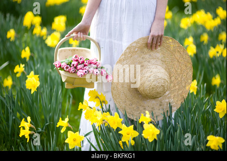 Frau trägt ein weißes Kleid hält eine hölzerne Trug der rosa Tulpen in einem Feld der gelben Narzissen und Stroh Sonnenhut Stockfoto