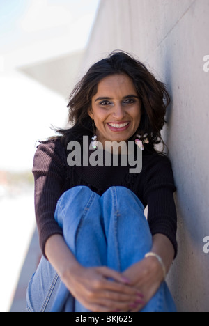 Eine schöne junge (Ost-) indische Frau sitzt und stützt sich auf eine helle farbige Wand - sie lächelt. Stockfoto