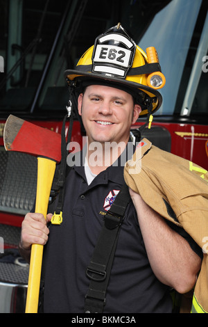 Fröhlicher Feuerwehrmann, der in Feuerwehruniform vor einem Feuerwehrauto steht Eine Axt halten und lächeln Stockfoto