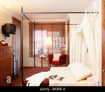 Weiße Vorhänge auf einfache Metall Himmelbett im Schlafzimmer mit Lattenrost aus Holz Rollo am Fenster und Fernseher an der Wand montiert Stockfoto