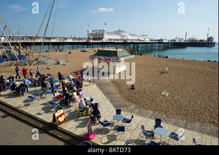 Leute sitzen an Tischen und Stühlen in einem Straßencafé auf Brighton Seafront, Brighton Pier im Hintergrund Stockfoto