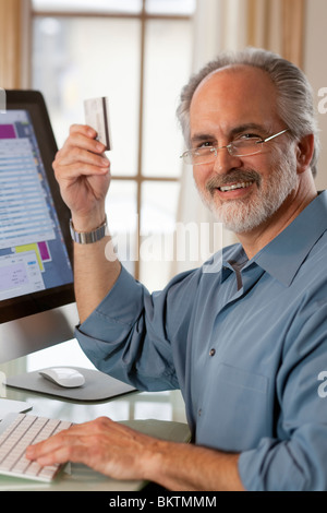 Ein Geschäftsmann sitzt vor einem Computer-Monitor und hält eine Kreditkarte. Er lächelt in die Kamera. Vertikale erschossen. Stockfoto