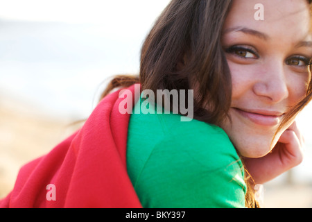 Weibliche in ihrem frühen 20er Jahren eingewickelt in eine rote Decke lächelt wie sie die Kamera am Strand sieht. Stockfoto