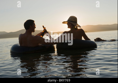 Ein junges Paar entspannen Sie bei einem kühlen Drink und Schwimmen in einem See bei Sonnenuntergang in Schläuche. Stockfoto