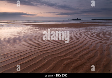 Wellen im Sand mit Dunstanburgh Castle in der Ferne im Morgengrauen Stockfoto