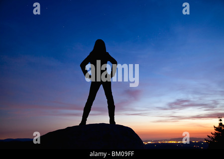 Silhouette einer Frau in der Nacht mit Blick auf die Lichter der Stadt auf einem Felsen steht Stockfoto