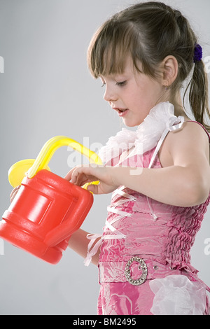 schöne Mädchen im rosa Kleid schaut eine rote Spielzeug Gießkanne auf grauem Hintergrund Stockfoto