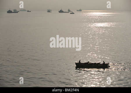 Ein Fischerboot im Hafen von Mumbai bei Sonnenaufgang mit Frachtschiffen im Morgennebel am fernen Horizont. Mumbai, Indien Stockfoto