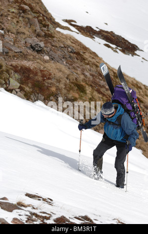 Ein Mann mit einem schweren Rucksack mit Skier geschnallt darauf klettern ein Schneehang mit Skistöcke für Unterstützung Stockfoto