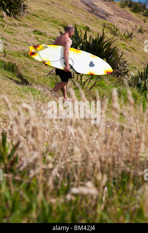 Eine Surfer trägt sein Board an den Strand. Muriwai Beach, Auckland, Nordinsel, Neuseeland Stockfoto