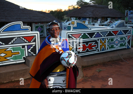 Eine Frau mit traditionellen Ndebele Stamm Farben Stand mit einem Fußball in einem kulturellen Dorf in Mpumalanga, Südafrika Stockfoto