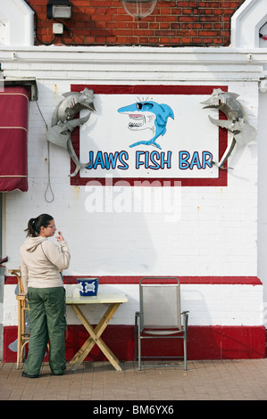 Eine weibliche Erwachsene Raucher unten ein Fisch Bar Zeichen Darstellung eine Hai-Cartoon-Figur auf Brighton Vorderseite Sussex UK Stockfoto