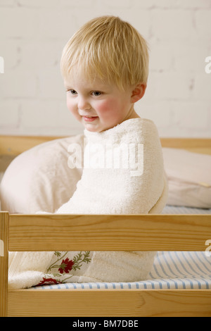 Ein glückliches Baby Junge eingewickelt in ein Handtuch sitzen auf einem Bett Stockfoto