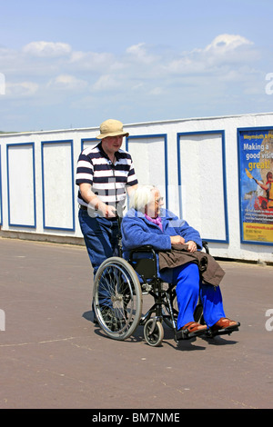 Ältere behinderte Frau sitzt im Rollstuhl und ist Beingpushed auf Weymouth Strandpromenade genießen den Frühsommer Sonnenschein Stockfoto