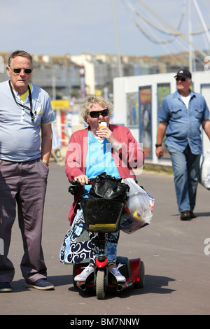 Ältere behinderte Frau reitet eine tragbare Mobilität Scooter Weymouth Strandpromenade entlang und genießt die Freude ein Eis Stockfoto