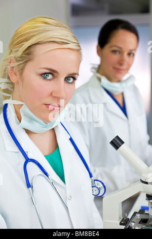 Eine blonde medizinische oder wissenschaftliche Forscherin oder Arzt mit ihrem Mikroskop im Labor mit ihrer Kollegin Stockfoto
