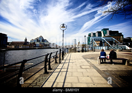 Zwei Personen sitzen auf einer Bank auf den North Quays von Dublin Docklands, allgemein bekannt als die Irish Financial Services Centre (IFSC) unter blauem Himmel. Stockfoto